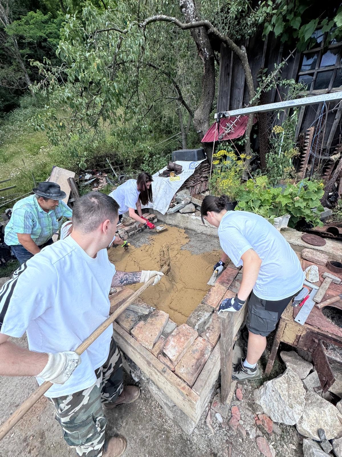 Tineri și un localnic lucrând cu mortar și cărămidă veche la baza unui cuptor tradițional