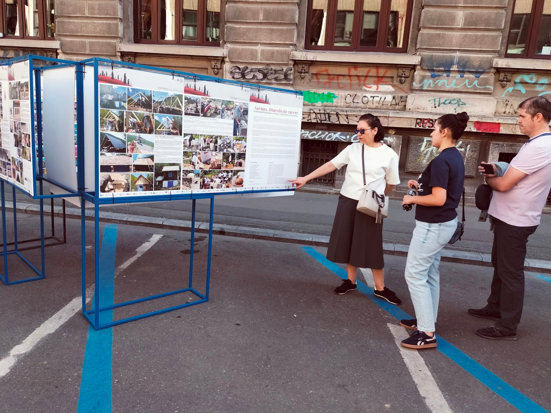 Trei persoane discutând în fața unui panou de expoziție cu fotografii despre patrimoniul rural